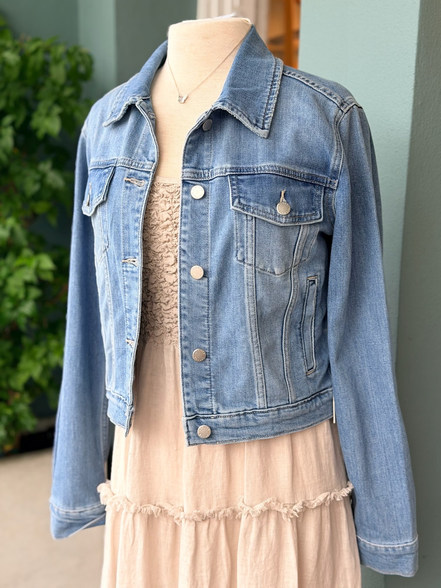 Maddy Medium Wash Denim Jacket front view. Styled over a beige dress with a silver butterfly necklace