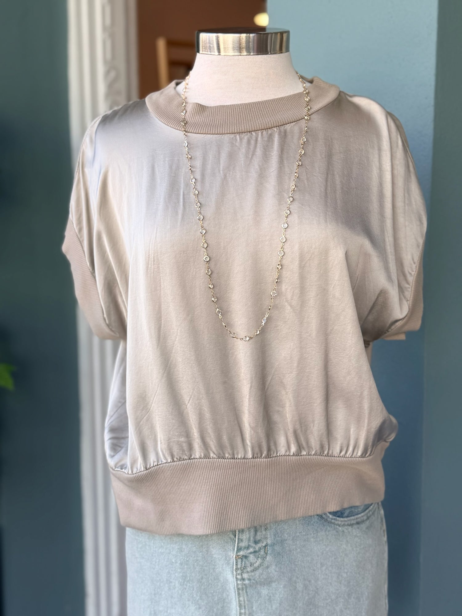 Mannequin wearing a beige blouse with a necklace against a blue wall.