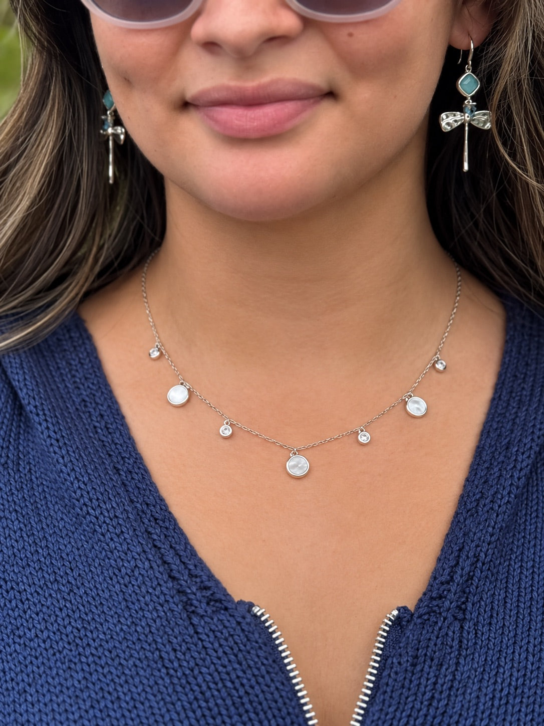 Close-up of the Melody Silver Necklace featuring a delicate silver-tone chain with round white mother-of-pearl inspired charms and small sparkling accents, modeled with a navy knit sweater.