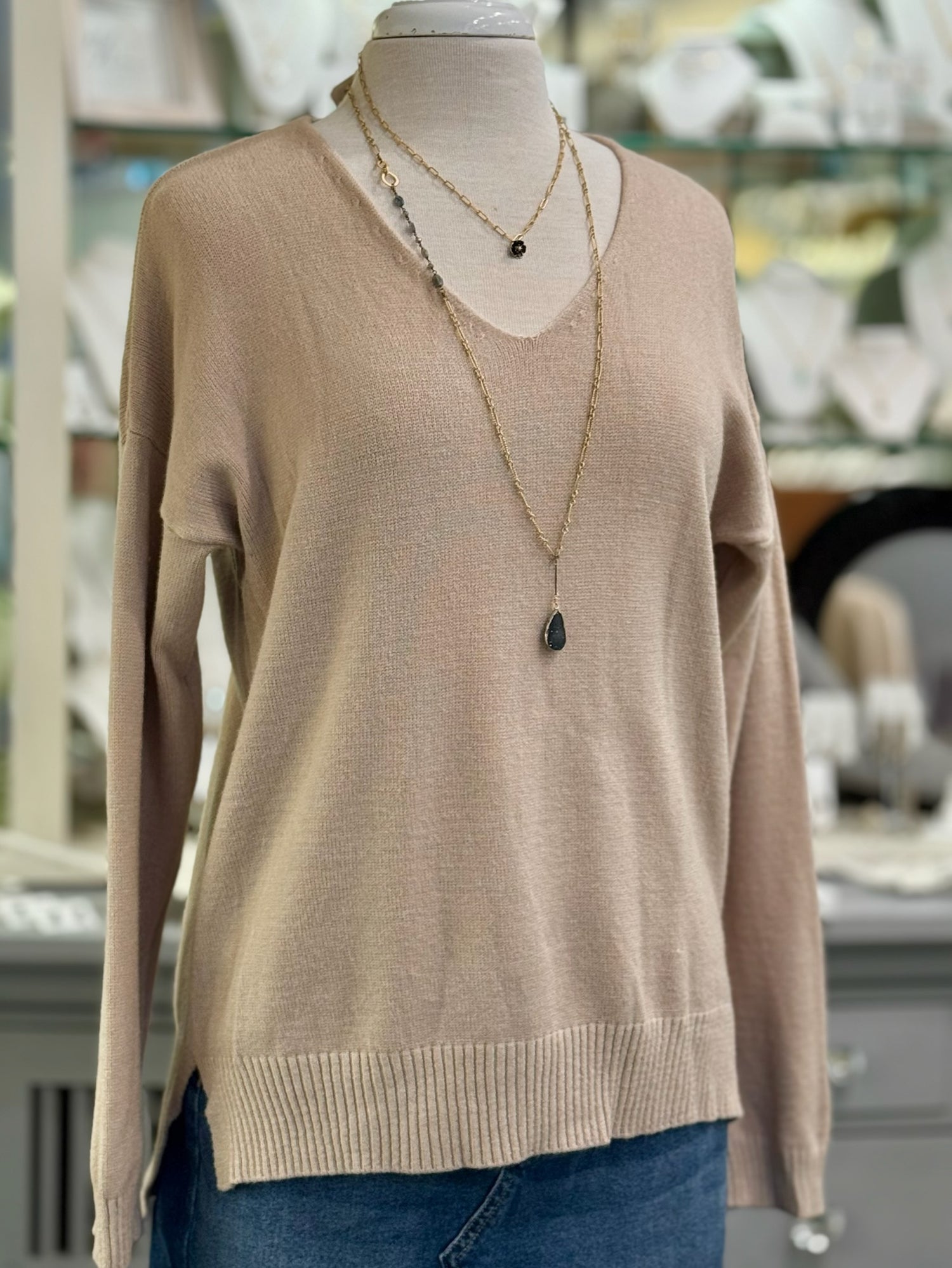 Blush taupe knit V-neck sweater on a mannequin, featuring a relaxed fit with ribbed hem and side slits, layered with gold necklaces.