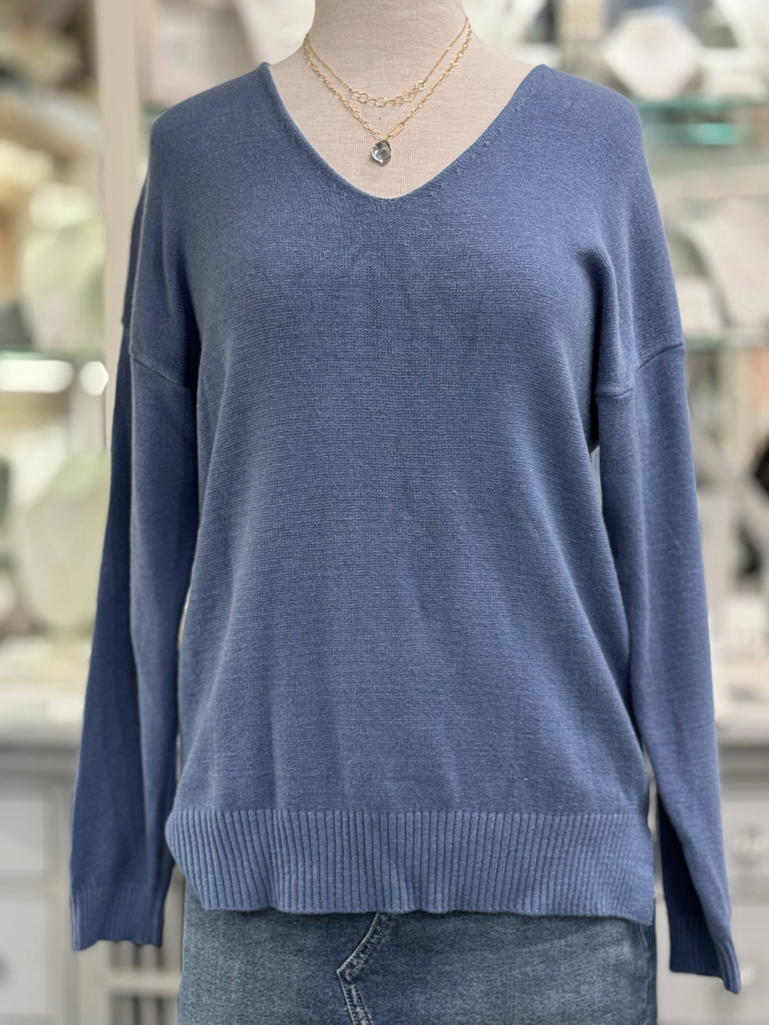 Dusty blue V-neck knit sweater on a mannequin, styled with gold chain necklaces and denim, featuring drop shoulders and wide ribbed hem.