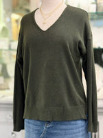 Olive green V-neck sweater with relaxed fit, drop shoulders, and wide ribbed hem, paired with gold chain necklaces and denim skirt.