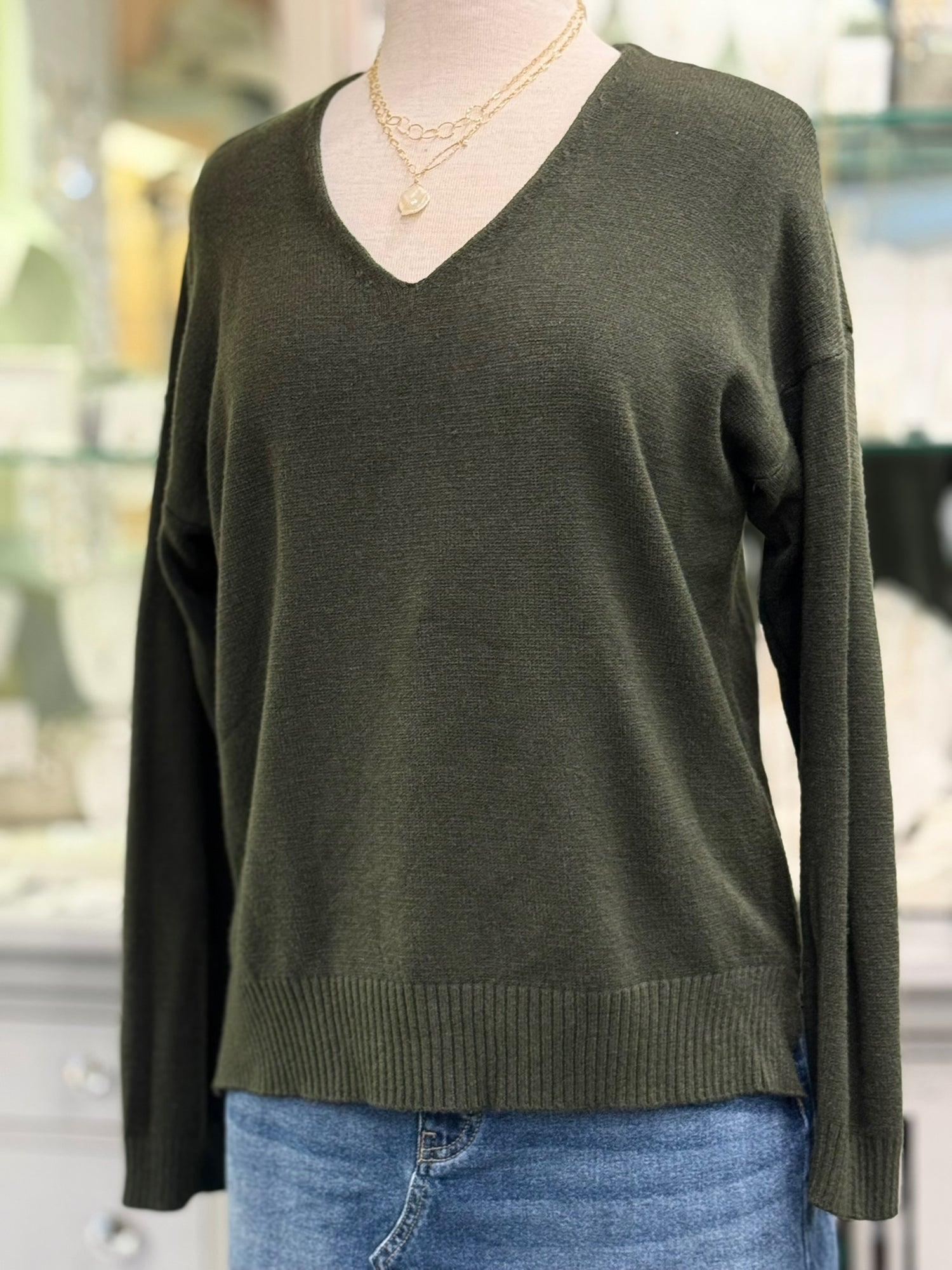 Olive green V-neck sweater with relaxed fit, drop shoulders, and wide ribbed hem, paired with gold chain necklaces and denim skirt.