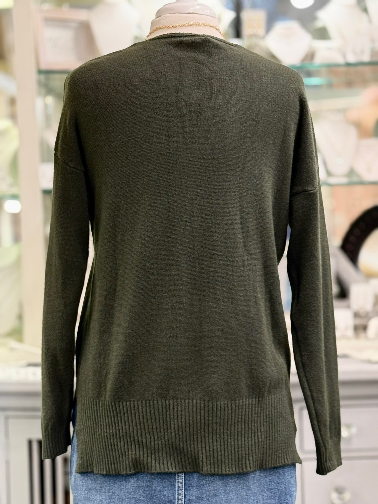 Back view of olive green knit sweater with long sleeves and ribbed hem, styled on a mannequin with denim.