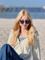 Woman wearing sunglasses oatmeal zip hoodie, sitting on a sandy beach with water and pier in the background
