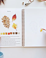 Open Autumn Watercolor Workbook showing leaf painting tutorials with watercolor steps and sketches, hand painting yellow and red autumn leaves.
