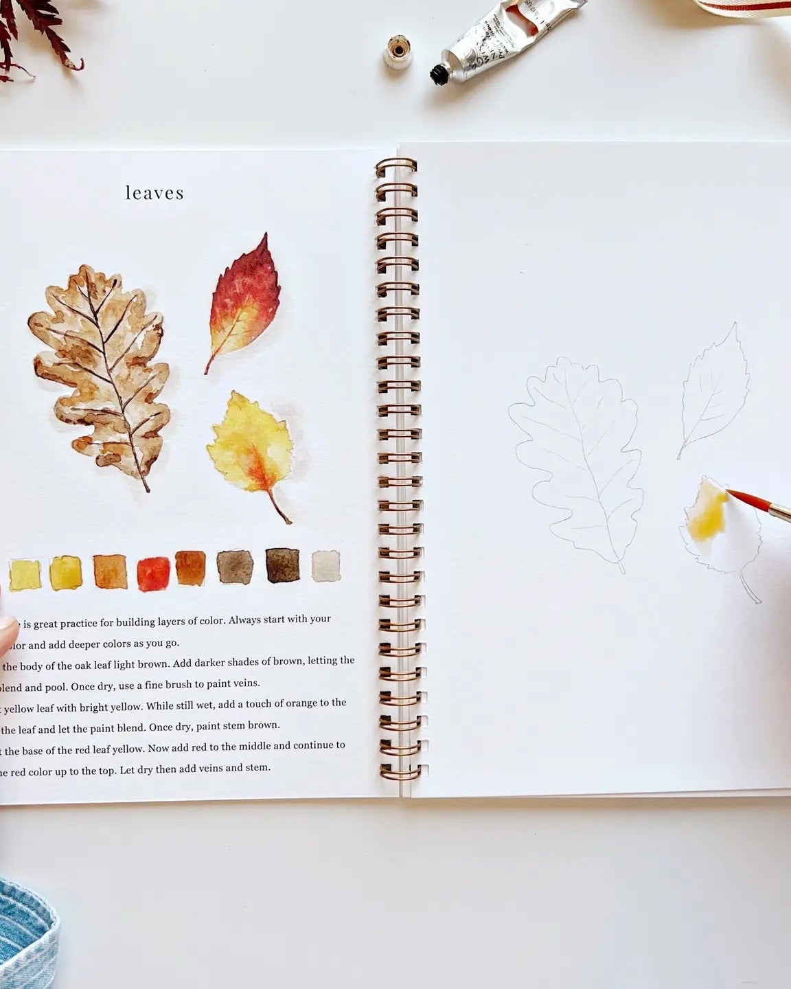 Open Autumn Watercolor Workbook showing leaf painting tutorials with watercolor steps and sketches, hand painting yellow and red autumn leaves.