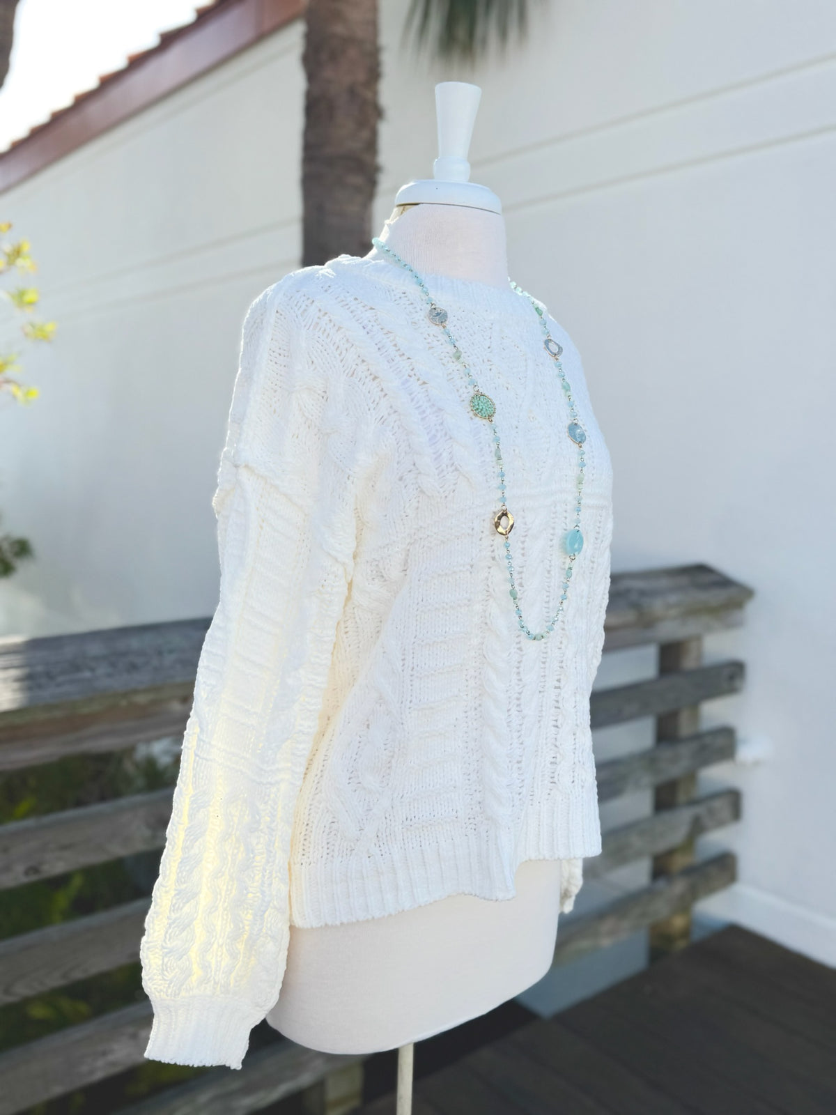 Side view of Cable Knit sweater in ivory on a mannequin outdoors with a long bright blue necklace.