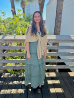 Woman in a green dress with a gold shawl standing on a wooden deck with trees and a building in the background.
