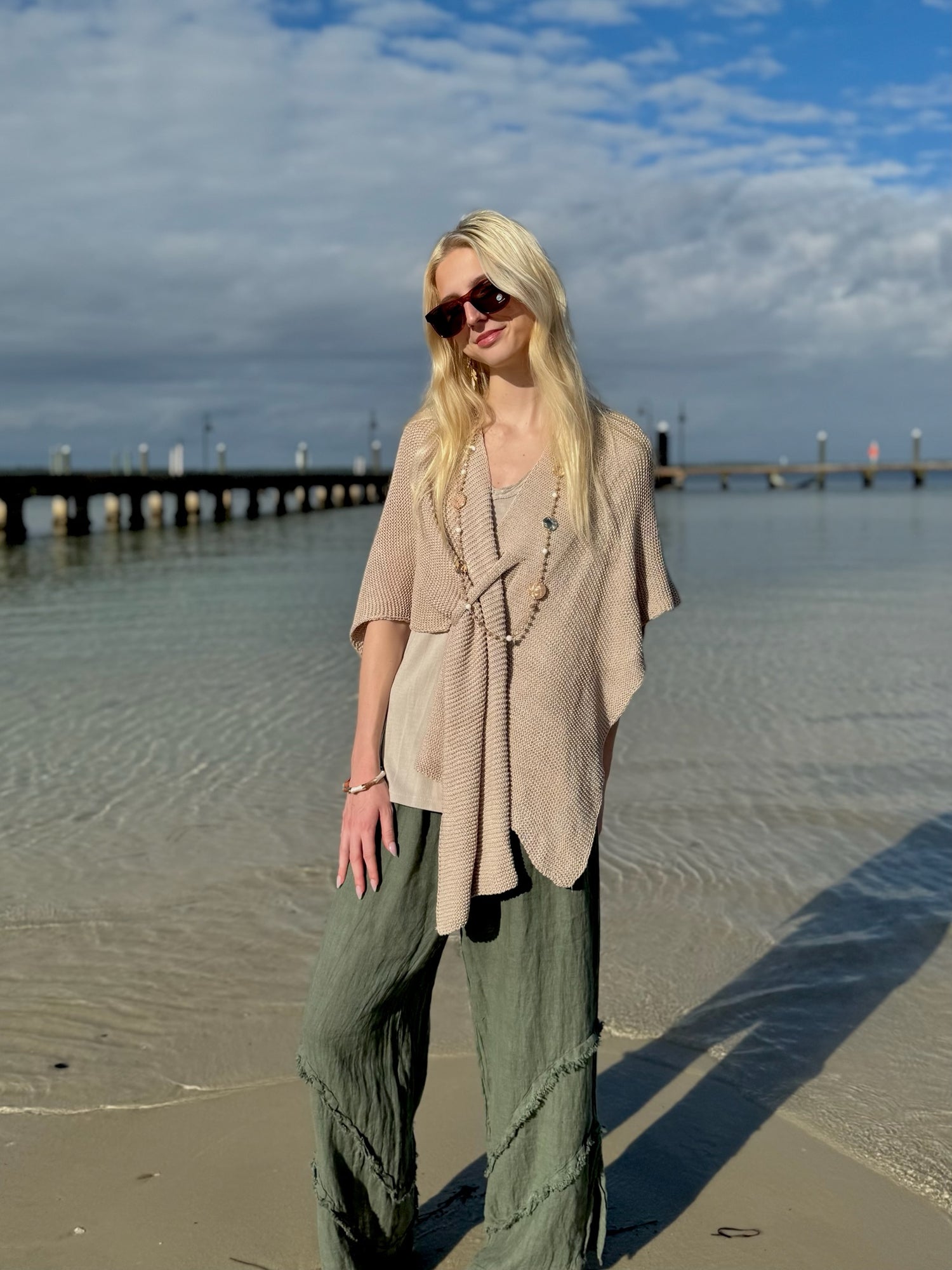 Model standing at the shoreline wearing a beige knit wrap poncho and olive wide-leg linen pants, with the ocean and pier in the background under a partly cloudy sky.