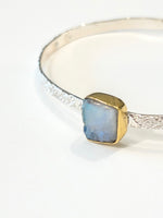A silver cuff bracelet featuring a rough cut Moonstone gemstone. Moonstone gemstone is set in gold bezel on textured sterling silver cuff