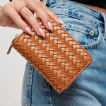 A tan woven leather zip-around wallet held by a person's hand, positioned over blue denim jeans.
