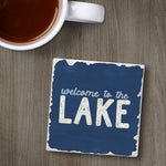 Coaster with 'welcome to the LAKE' text next to a cup of coffee on a wooden surface