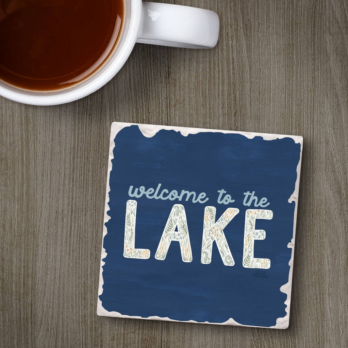 Coaster with 'welcome to the LAKE' text next to a cup of coffee on a wooden surface