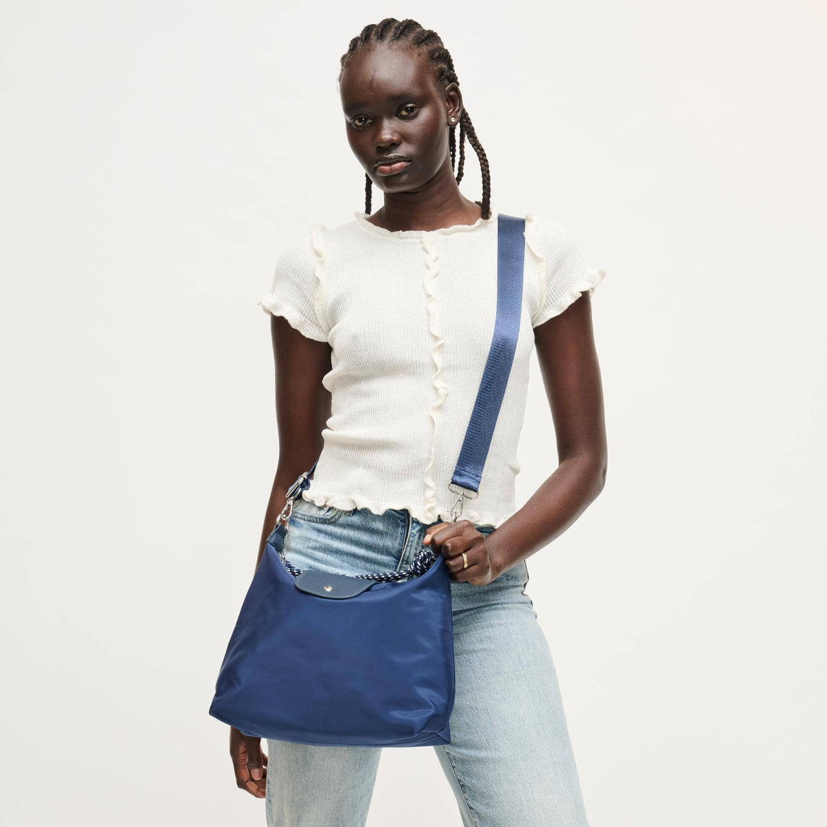 A model wearing a white ruffle-trimmed short-sleeve top and light-wash jeans carries a blue nylon shoulder bag with a wide strap and silver-tone hardware against a plain off-white background.