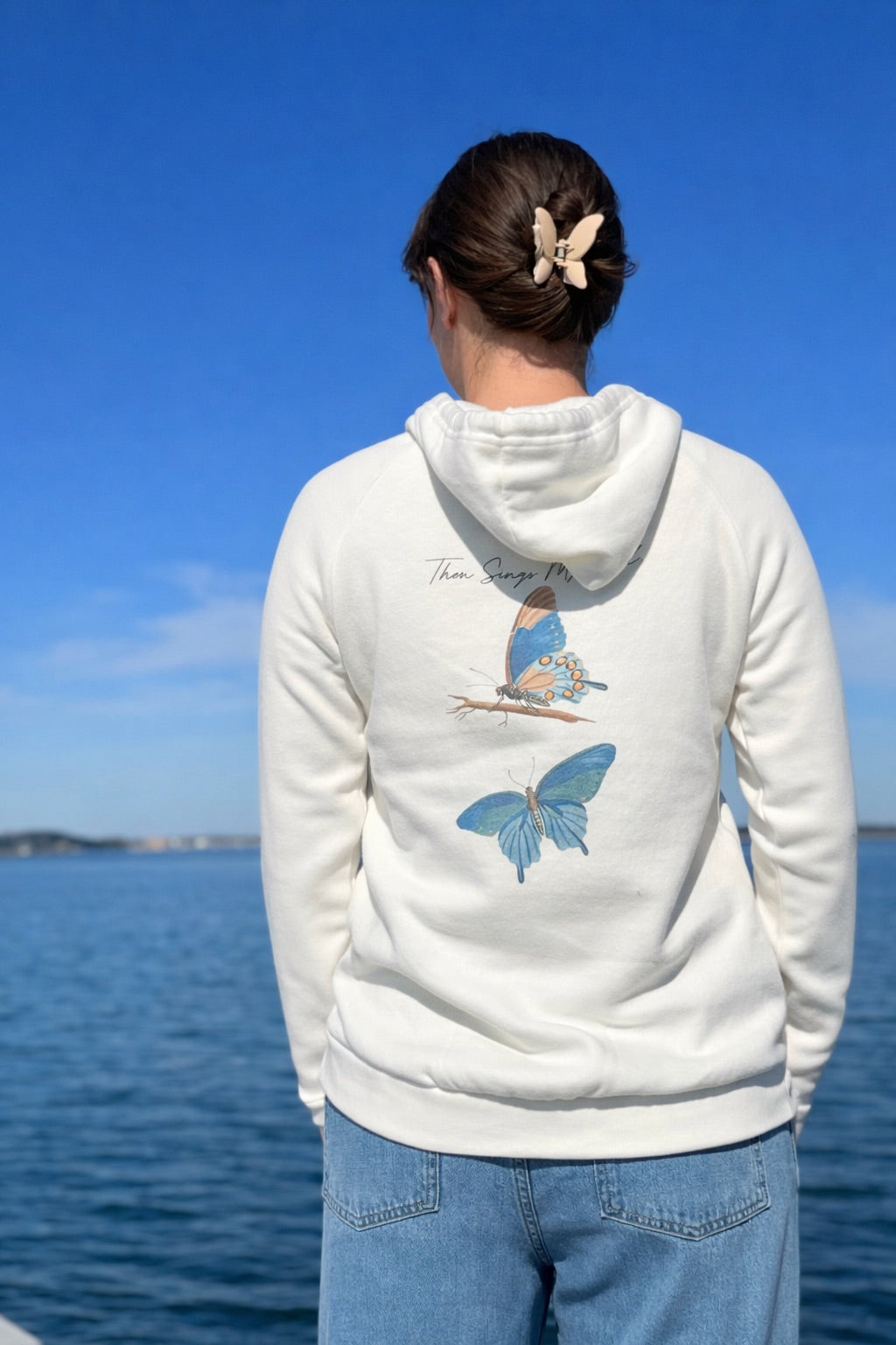 Person wearing an Ivory hoodie with butterfly designs by a body of water