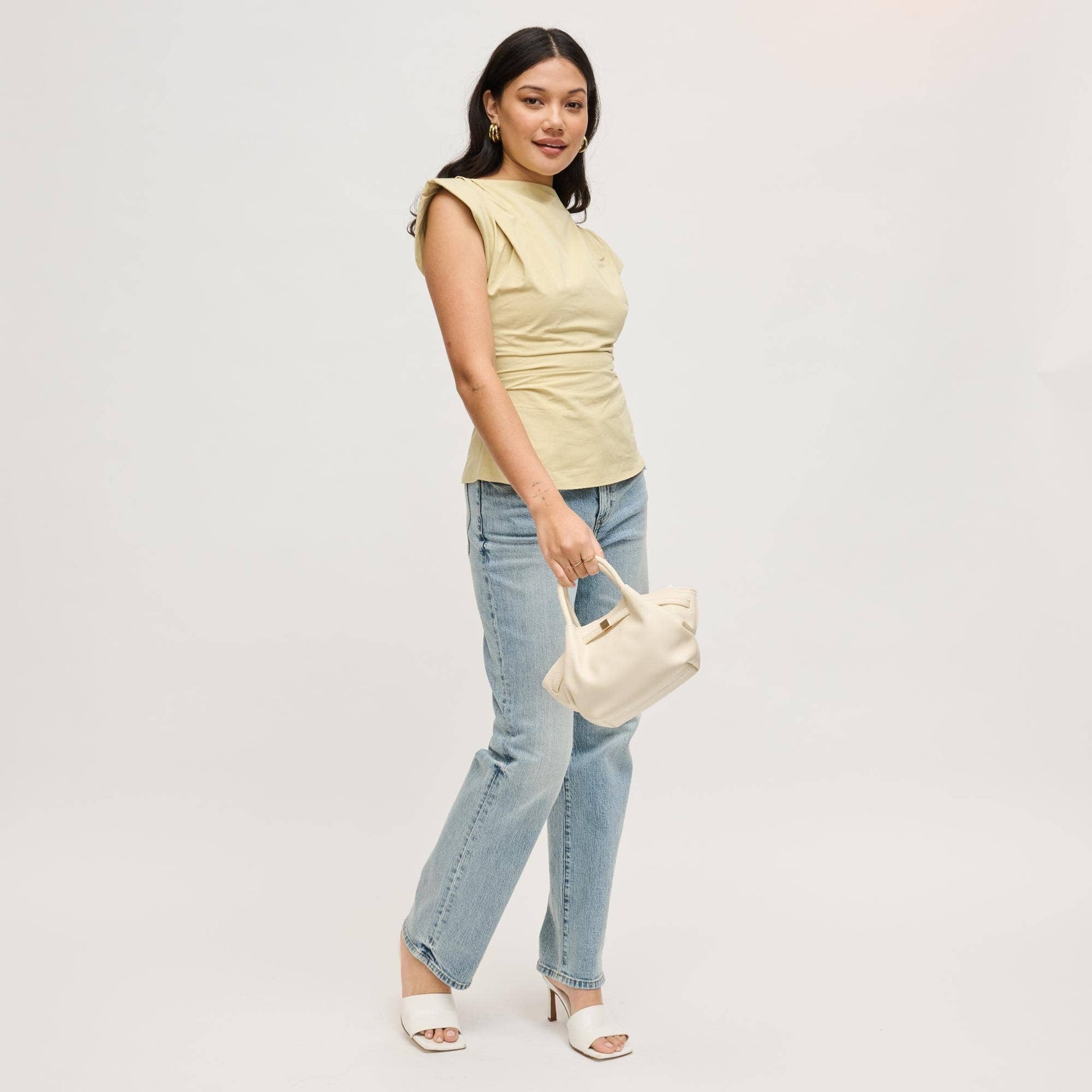 Model carrying the Selena Crossbody in cream by its rolled top handles, featuring soft vegan leather, a gathered silhouette, and polished gold-tone hardware.