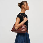 A woman in a black top and blue skirt is shown in profile carrying a brown woven leather shoulder bag with a braided strap against a plain light gray studio background.