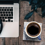 Top-down view of a workspace scene with part of a silver laptop, a cup of black coffee on a patterned saucer, and dark leaves on a rustic wooden table.
