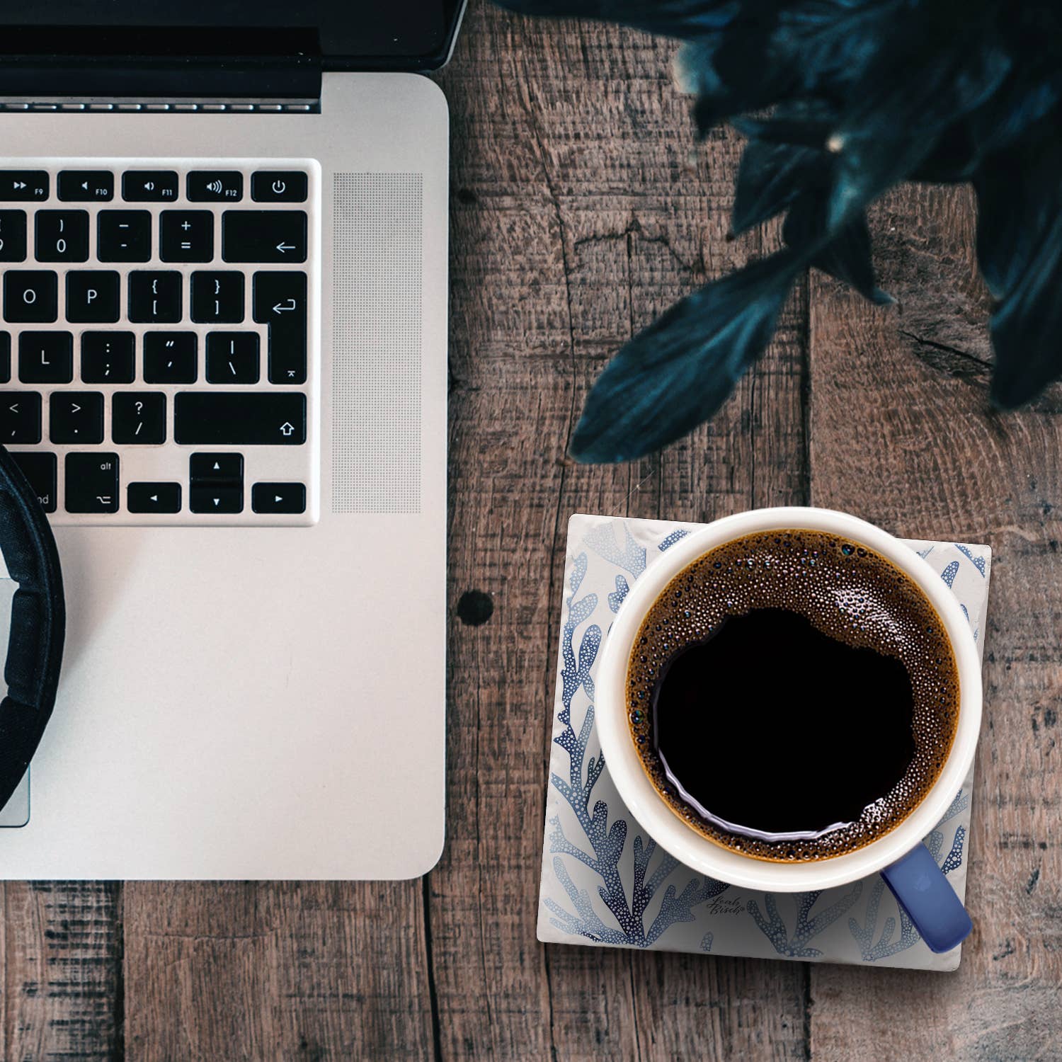 Top-down view of a workspace scene with part of a silver laptop, a cup of black coffee on a patterned saucer, and dark leaves on a rustic wooden table.