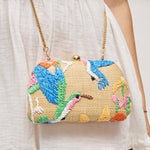 A close-up of a woven clutch handbag with colorful embroidered birds and floral motifs, hanging from a gold chain strap against a white sleeveless dress.