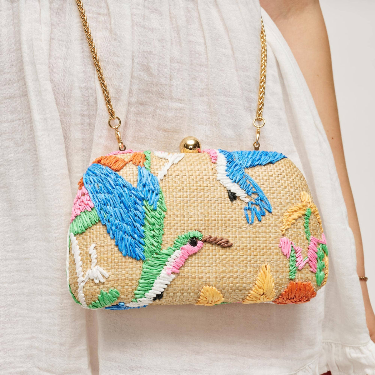 A close-up of a woven clutch handbag with colorful embroidered birds and floral motifs, hanging from a gold chain strap against a white sleeveless dress.
