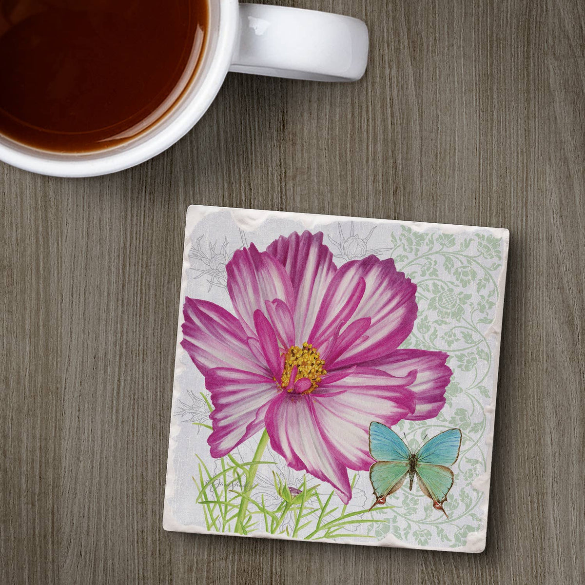 A square ceramic coaster with a printed design of a pink and white cosmos flower, green stems, and a light blue butterfly, shown on a gray wood-grain tabletop next to a white mug filled with dark coffee.