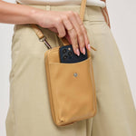 A tan nylon crossbody phone pouch worn on a model's hip, with a hand sliding a black smartphone into the front pocket.