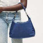 A person wearing light blue jeans and a white top holds a blue nylon shoulder bag with a braided blue-and-white strap and silver hardware against a plain light background.