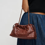 A brown woven leather shoulder bag with a gold-tone chain strap, carried by a model wearing a black top and blue skirt against a light gray background.