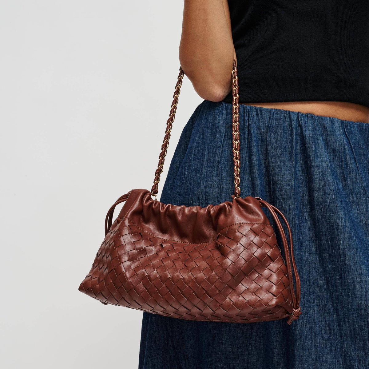 A brown woven leather shoulder bag with a gold-tone chain strap, carried by a model wearing a black top and blue skirt against a light gray background.