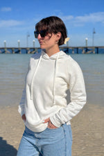 Woman wearing an Ivory hoodie and sunglasses on a beach with a pier in the background