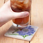 A hand holds a glass of iced coffee over a square ceramic coaster printed with a blue flower and hummingbird design, set on a light wood tabletop.