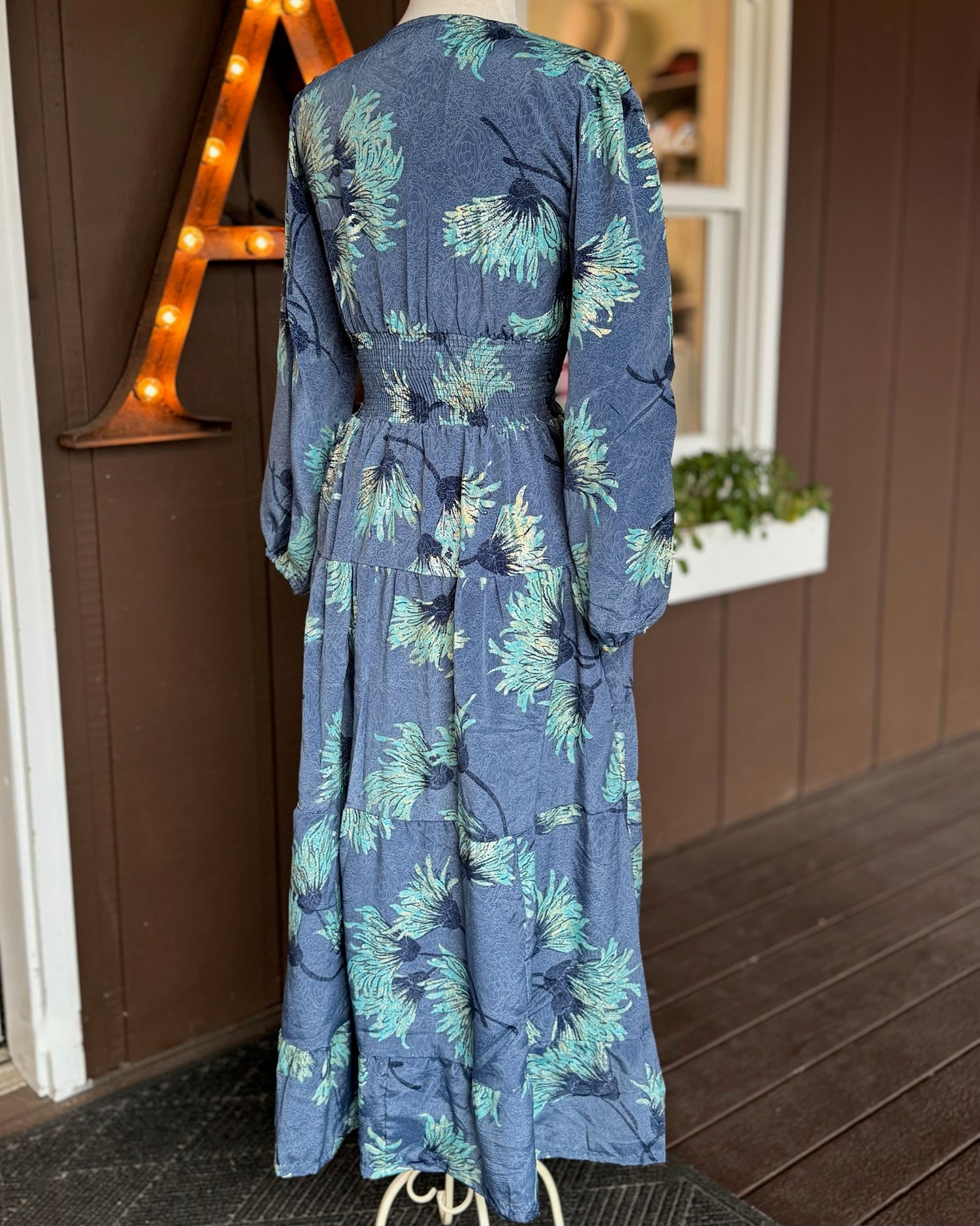 Back view of blue floral maxi dress featuring aqua and gold print, smocked waist, and flowing tiered skirt