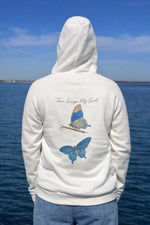 Person wearing an Ivory hoodie with butterfly designs and text by a body of water.