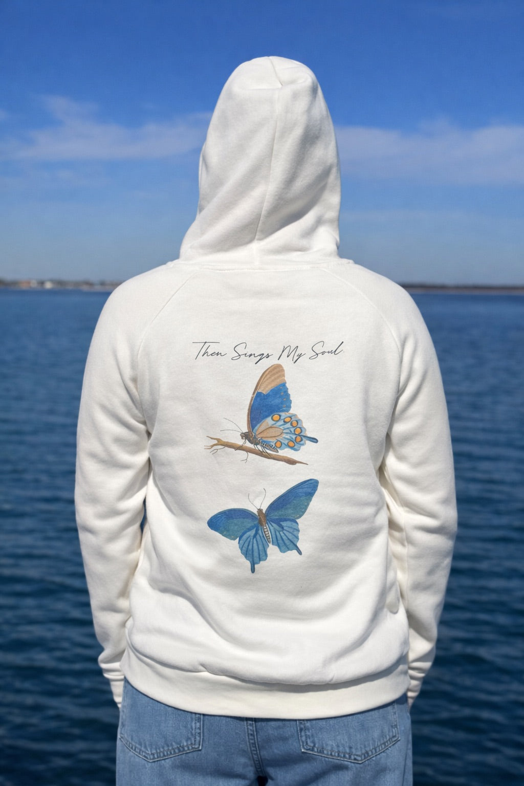 Person wearing an Ivory hoodie with butterfly designs and text by a body of water.