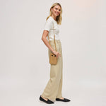 A woman modeling a white short-sleeve knit top, beige wide-leg trousers, and black flats, carrying a small tan crossbody bag against a plain light gray studio background.