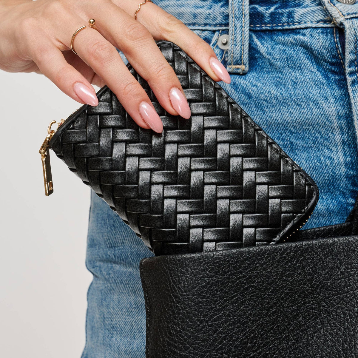 A close-up of a person's hand with pink manicured nails holding a black woven leather zip pouch next to a black textured leather bag, set against blue denim jeans.