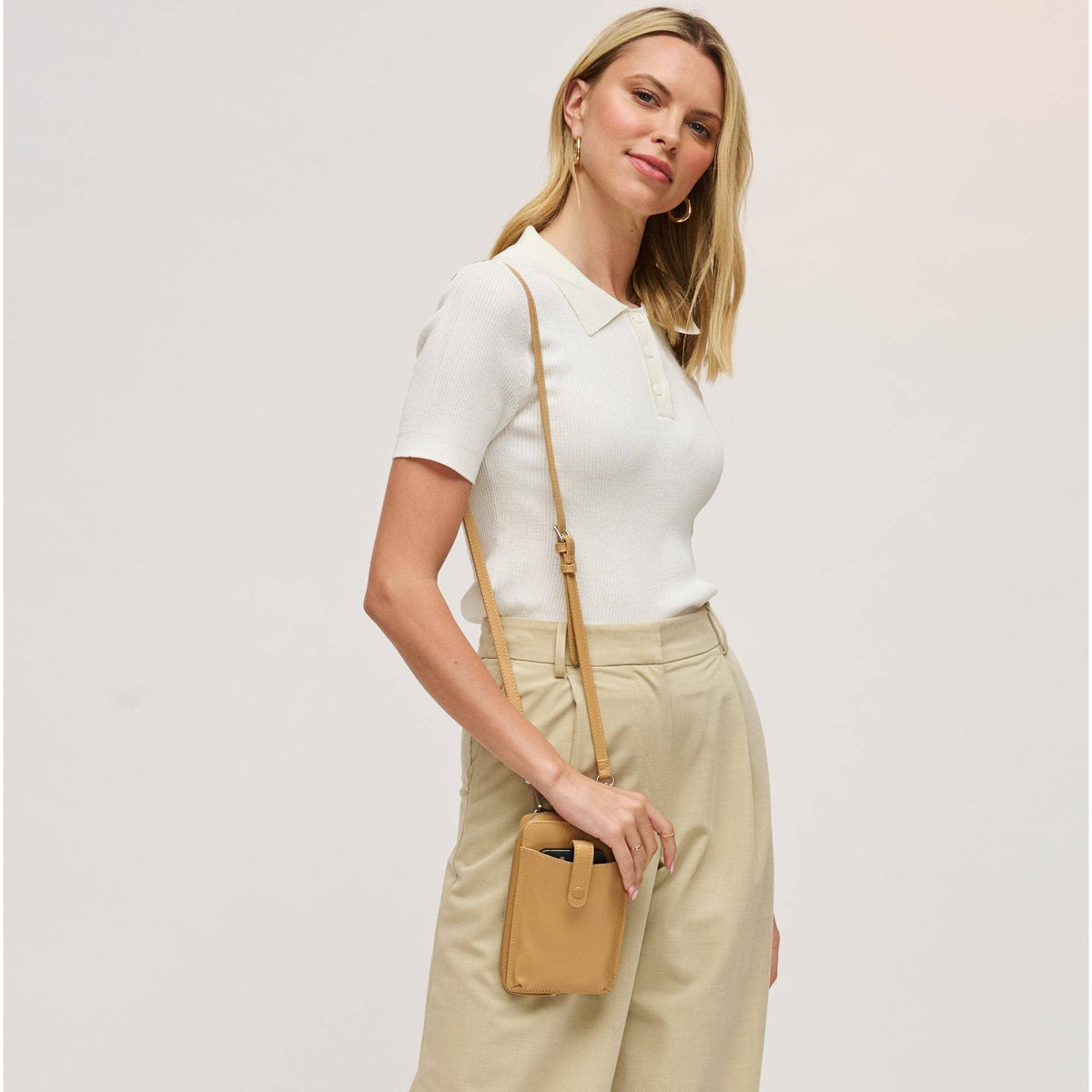 A woman wearing a white short-sleeve polo shirt and beige trousers, carrying a small tan crossbody bag with a matching strap against a light gray studio background.