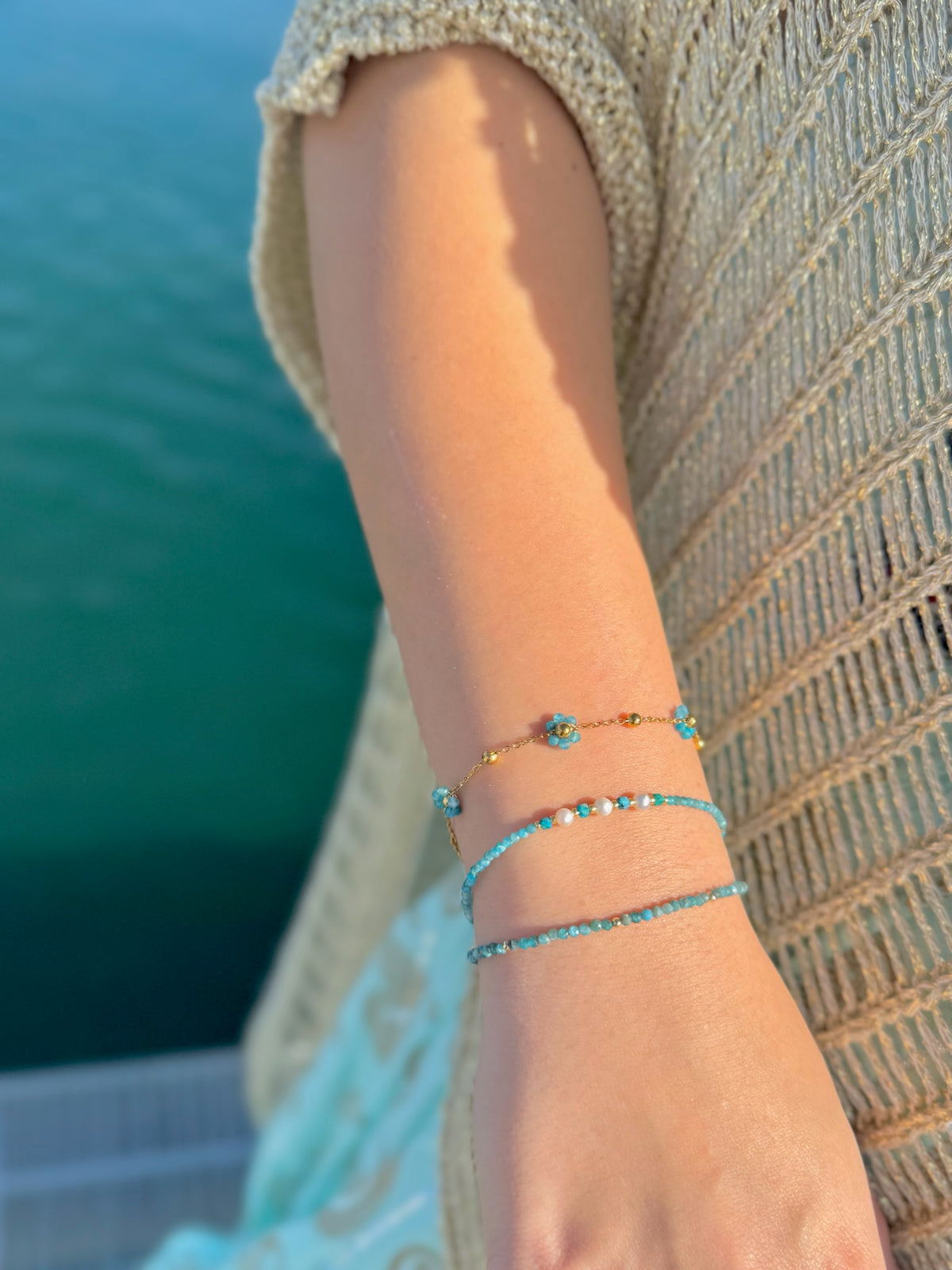 Blue apatite flower bracelet stacked with two blue-green beaded bracelets on a wrist outdoors beside the water.