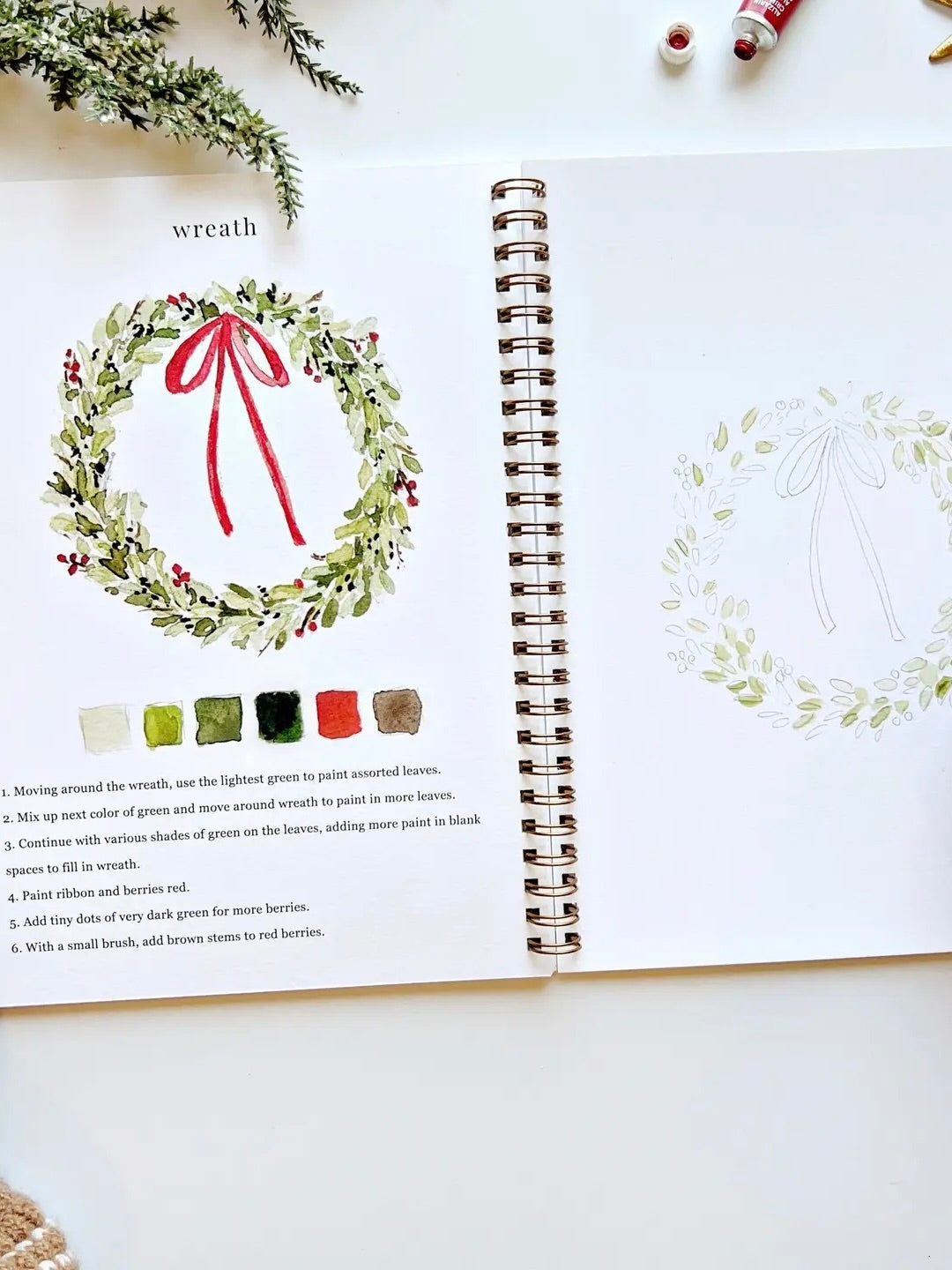 Christmas Watercolor Workbook open to wreath painting page with step-by-step instructions, color palette, and outline sketch by Emily Lex