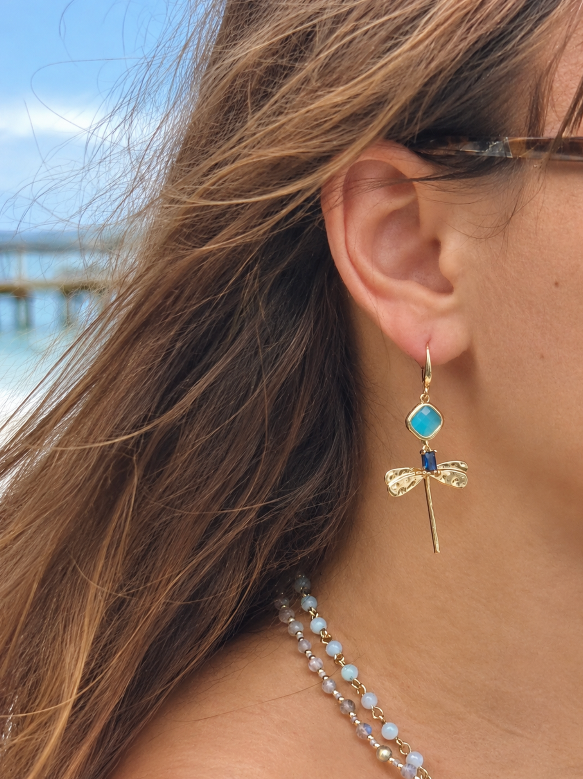 Close-up of a Lila gold-tone dragonfly earring worn on the ear, showing its aqua stone, navy accent and textured wings.