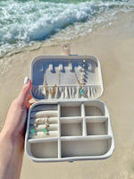 A travel jewelry organizer in a creamy white color, with multiple compartments containing various pieces of jewelry, held open by a hand against a beach and ocean backdrop.