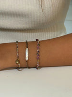 Three delicate beaded bracelets stacked on a wrist, featuring multicolored gemstones, gold accents, a small gold charm, and a vertical baroque pearl centerpiece against a neutral background.
