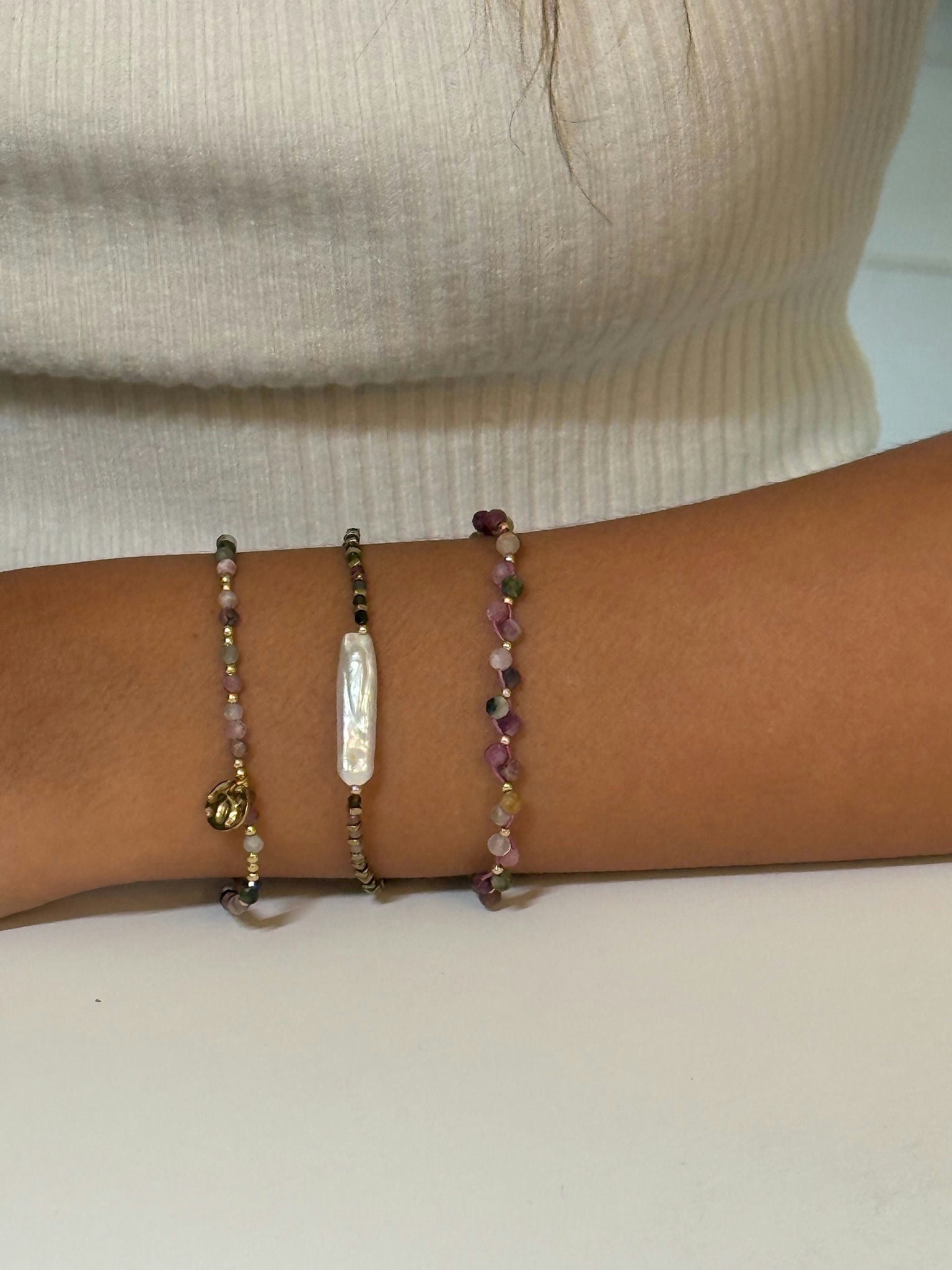 Three delicate beaded bracelets stacked on a wrist, featuring multicolored gemstones, gold accents, a small gold charm, and a vertical baroque pearl centerpiece against a neutral background.
