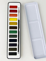Emily Lex watercolor paint set open to reveal twelve color pans and a round brush, with the white tin lid shown beside as a mixing palette.