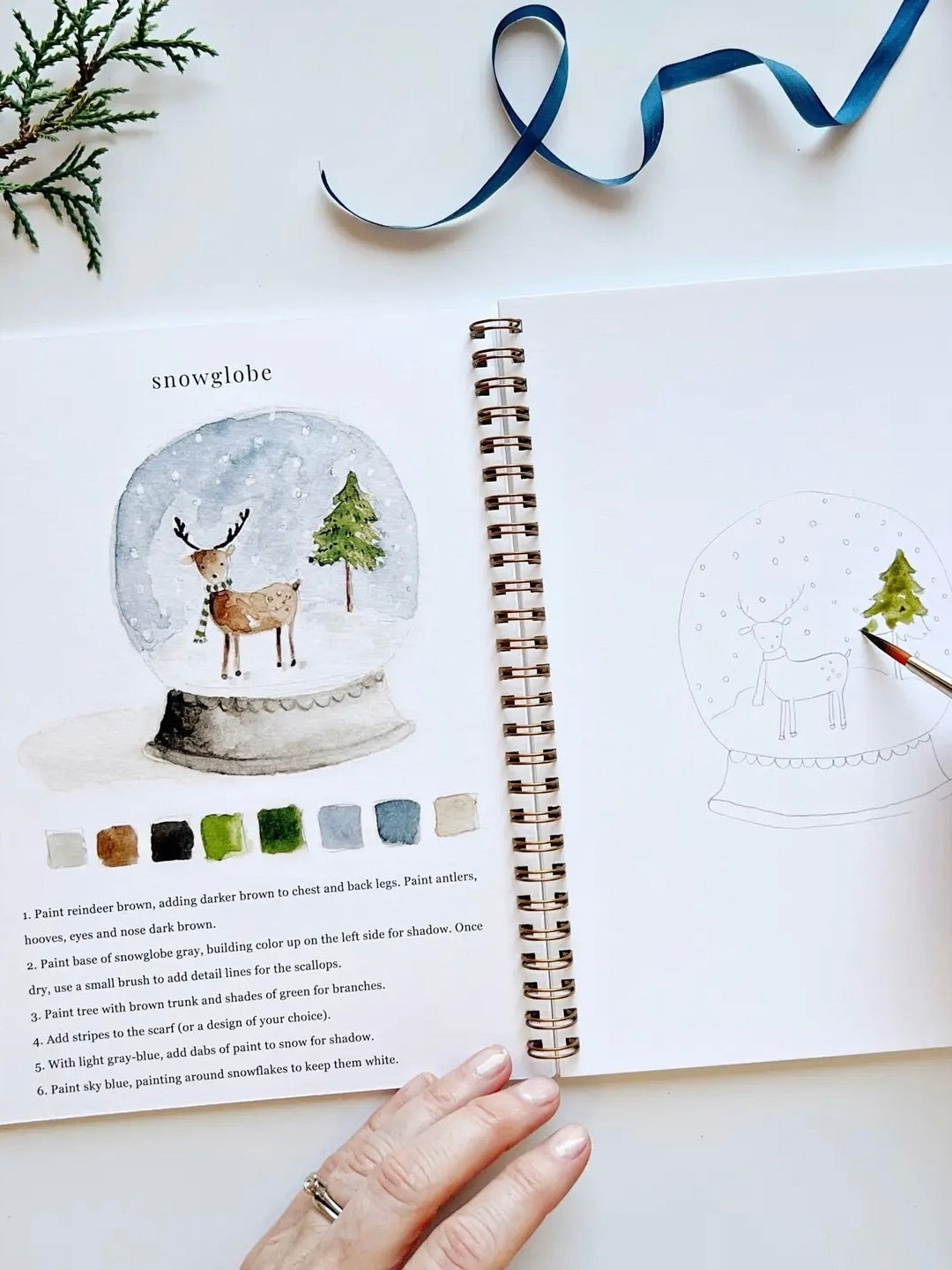 Winter Watercolor Workbook open to snowglobe painting page with step-by-step instructions, color palette, and outline sketch by Emily Lex
